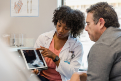 The mid adult female orthopedic surgeon points to the x-ray of the patient's foot on digital tablet as she explains treatment options to the mature adult man.
